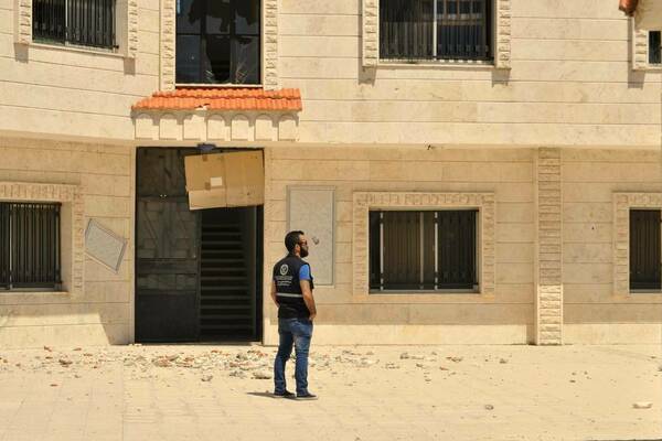 Man in dark vest and jeans looks right at a light-colored building with a broken upper window and ground debris.
